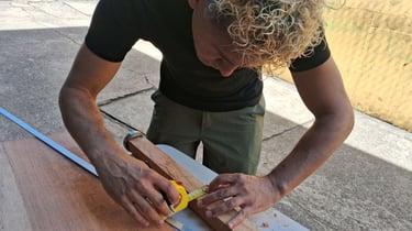 A carpenter with curly blonde hair uses a yellow measuring tape on a wooden plank for a DIY project.