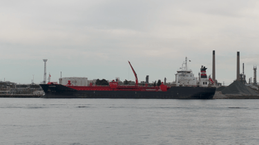 A blue hulled tanker ship with a white superstructure and red deck, docked at a refinery.