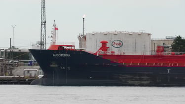 The blue bow and red deck of a tanker ship in front of large oil tanks at a refinery.