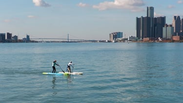 2 men paddleboarding with the Ambassador Bridge and Detroit and Windsor skylines in the background.