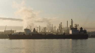 A blue tanker ship moored in front of a refinery with steam in the background.