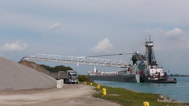 A semi-truck drives under the boom of a self-unloading barge, unloading at a stone yard.