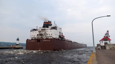 The stern of a huge red ship passing between 2 lighthouses at the entrance of the Duluth Ship Canal.