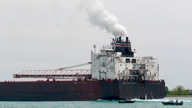 The aft section of a huge ship with a plume of steam exhaust, passing 2 small boats.