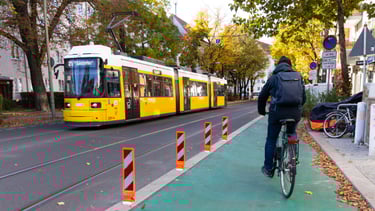 Bild von einer Straße mit grünem Fahrradweg. Auf der anderen Straßenseite fährt eine Straßenbahn.