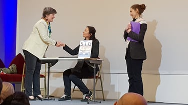 Sur scène une femme debout serre la main sous le regard d'une troisème forum accueil usager