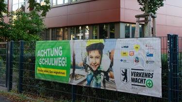 Foto von einerm roten Schulgebäude, an dem Zaun davor ist ein Plakat zur Schulwegsicherheit.