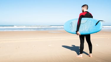 En homme en combinaison de surf, avec une planche bleu sur une plage
