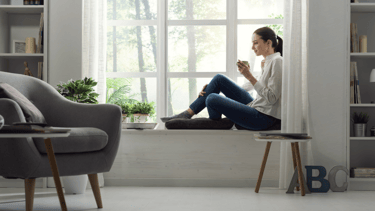 Habitación relajante, luminosa, pintada de blanco, con sillón gris y mujer sentada junto al ventanal