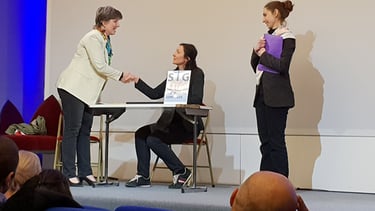 Sur scène une femme debout serre la main sous le regard d'une troisème forum accueil usager