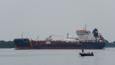 A man is standing up fishing in a small boat as a tanker ship passes behind him.