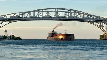 Sun shines on the bow of a huge red freighter, with a steam exhaust plume, passing under a bridge.
