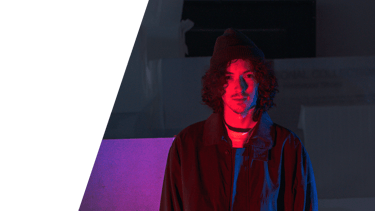 A dark portrait photographic image of In Videcolour lead-songwriter Sam Auckland. Sam is standing, red and blue light on him.