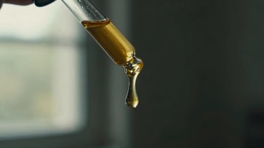 Macro close-up of a glass apothecary dropper releasing a single golden amber drop of kelulut honey, soft side-lit window sun, dark moody background