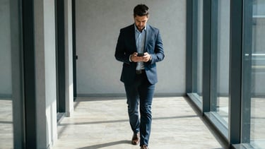 A sharp, high-angle photograph of a modern tech professional walking through a minimalist, sun-drenched corridor in a North American / US metropolitan office. They are looking at a smartphone with a focused expression. The color palette includes deep navy and light gray shadows with bright blue accents from the glass windows. Professional, clean, and dynamic atmosphere.