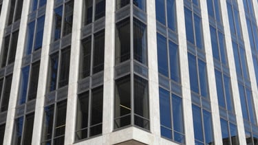 A sharp, professional architectural photograph of a modern steel and glass office building in a North American / US city. The shot is taken from a street-level perspective looking up. Lighting is bright daylight, highlighting clean white concrete and navy blue tinted glass. The composition emphasizes strength and reliability.