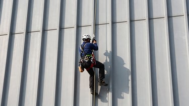 Rope-access technician in full harness and helmet working on a vertical steel industrial facade, rigging lines taut, hands gripping a tool at a structural connection point, hard daylight shadows on the structure surface, wide framing showing height and scale