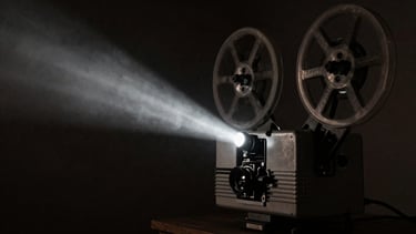 Dark cinematic photography, low-key chiaroscuro lighting, deep shadows, a vintage film projector casting a sharp beam of light through smoke, charcoal tones, 35mm grain.