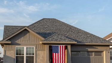 A clean, eye-level photograph of a residential house in Colorado Springs with a brand new asphalt shingle roof in a medium gray color. The house has a well-maintained lawn and a North American / US flag proudly displayed by the front door. The sky is a bright blue with soft morning light, and the overall feel is one of quality craftsmanship and patriotic pride.