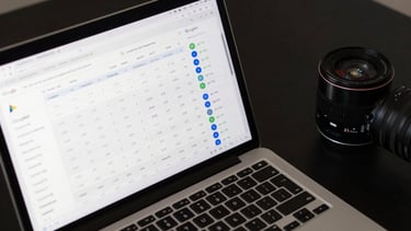 Close-up overhead of a laptop screen displaying a Google Ads performance dashboard with cost-per-conversion metrics, click data columns, and campaign status indicators under clean studio light on a dark desk surface