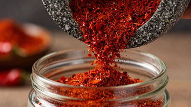 Macro shot of vibrant crimson red chili powder cascading from a stone mill into a clean glass jar, dramatic side-lighting, shallow depth of field.