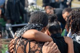 Mourners in black lace clothing embrace and comfort each other during an outdoor funeral service.
