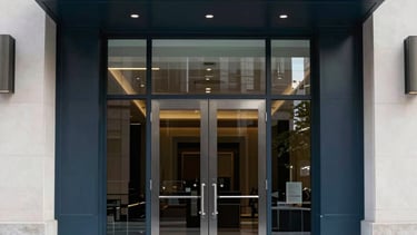 A professional architectural photograph of a high-end commercial building entrance in a North American business district. The shot features large, modern dark steel and glass double doors with polished hardware. The lighting is crisp, bright daylight, highlighting the clean, reliable aesthetic. The color palette incorporates deep blues and off-white tones.