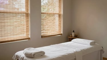 Serene, uncluttered treatment room with natural light filtering through bamboo blinds, a single massage table with clean white linens, minimalist organic decor, soft editorial style.
