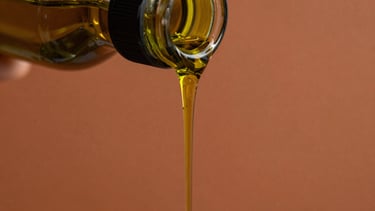 Macro close-up of golden-green olive oil slowly dripping from a dark glass bottle, catching high-contrast golden hour sunlight, rich terracotta background