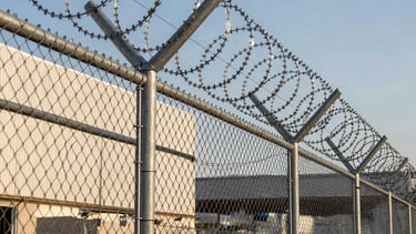 Detailed architectural photograph of a professional perimeter security installation in a South American industrial park. A perfectly tensioned galvanized silver chain-link fence with a concertina wire top reflects the afternoon sun. The background shows a clean modern warehouse building under a clear sky. Serious and robust atmosphere with sharp focus on metallic textures.