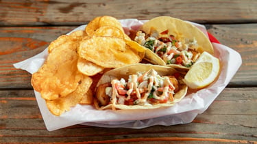 Fish tacos with cabbage, tomatoes, and sauce served with potato chips and a lemon wedge from Captain’s Galley