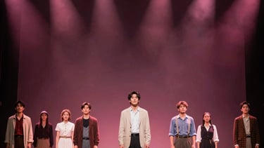 Full stage shot of a theater production with dynamic light cues featuring light pink and deep burgundy hues, focused on North American / US actors in a dramatic scene.