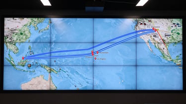 Wide panoramic GPS route overlay map on a large logistics control room display screen, active shipping lanes rendered as bright steel-blue lines crossing the Pacific and Indian Ocean, port nodes marked with precise coordinate pins at Singapore, Jakarta, Rotterdam, Shanghai, and Los Angeles, hard overhead studio lighting illuminating the screen surface, no people visible, geometric and orderly composition