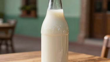 A professional, minimalist close-up photograph of a sleek glass bottle of fresh, white kefir resting on a sunlit wooden table in a traditional Latin American Mexican patio in Jalisco. The lighting is soft and golden, highlighting the smooth texture of the product. The color palette features sage green and cream tones.