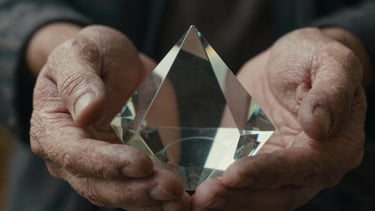High-contrast wide anamorphic cinematic still, extreme close-up of weathered hands holding a delicate vintage glass prism, moody tungsten light leak.