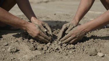 Wide panoramic shot of hands caked in dry river silt, scooping raw clay from a riverbed, harsh directional side-lighting, deep shadows, desaturated earth tones.