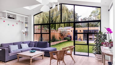 Modern living room featuring a purple sectional sofa and large black steel-framed windows overlooking a lush garden.