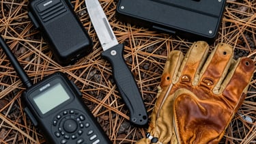 A flat lay photography of essential overlanding gear: a portable radio, a rugged knife, a satellite communicator, and a pair of leather gloves on a bed of dry pine needles and forest soil. Sharp focus, high contrast lighting.
