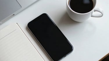 A top-down view of a designer's desk in a North American studio featuring a sleek smartphone, a cup of black coffee, and a minimalist notebook under bright, cool white light.