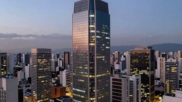 A twilight aerial photograph showing the illuminated skyline of São Paulo, focusing on a specific modern office tower. Clean, sharp lines and professional architectural lighting style.