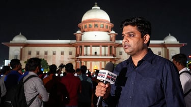 A high-contrast wide 16:9 shot of a live television news broadcast, a reporter holding an Aajeevak News microphone in front of a busy government building, dramatic press lighting, a subtle red live indicator in the corner.