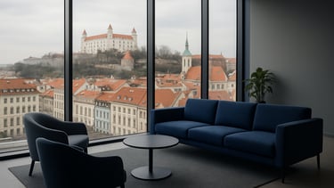 A crisp, high-end commercial photography shot of a modern office lounge in Bratislava, Slovakia. Through a floor-to-ceiling glass window, clean Central European city architecture is visible under soft overcast light. The interior features sleek furniture in midnight blue and muted grey textiles. A professional atmosphere reflecting synergy and reliability, with natural daylight and a minimalist composition.