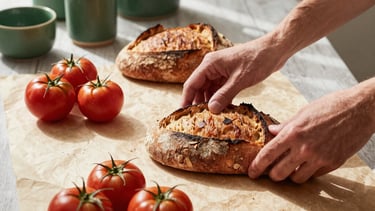 A high-end lifestyle photography shot of a content creator in a sunlit, Scandinavian-style studio in a Western European / North American city. They are styling a flat-lay of fresh sourdough bread and heritage tomatoes on a Crisp Parchment surface. The lighting is natural and soft, with accents of Matte Forest Green in the background ceramics. Professional, cozy, and sophisticated atmosphere.