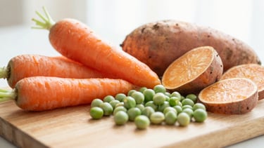 A close-up of fresh, vibrant organic vegetables (carrots, sweet potatoes, peas) on a wooden cutting board, diffused natural morning light, soft and warm, ready for baby food preparation, warm editorial photography.
