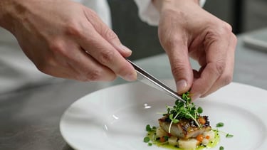 Candid motion-blurred close-up of a chef's hands using precision tweezers to place a micro-herb on a dish, clean stainless steel background, dramatic lighting.