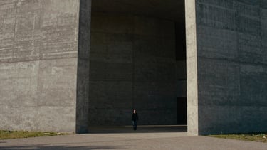 Cinematic film photography, moody natural light, high contrast, desaturated earth tones, wide anamorphic frame, 35mm grain, extreme wide shot of a colossal brutalist concrete structure with a tiny human figure standing in the shadows