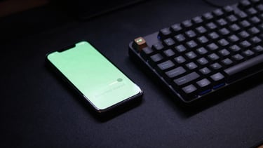High-angle desk setup at dusk. A sleek Android phone rests next to a mechanical keyboard, its screen casting a soft green glow on the matte black desk surface, moody tech photography
