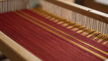 Macro close-up of a wooden loom with deep red and gold threads, soft warm side-light highlighting the physical texture of the weave, shallow depth of field
