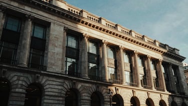 Low-key architectural shot of a historic London neoclassical facade blended with modern glass, dramatic chiaroscuro lighting, deep obsidian and gold tones, 35mm film grain.