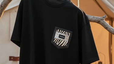 Black crewneck t-shirt with a patterned chest pocket hanging on a tree branch near a camping tent.